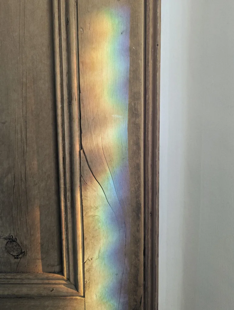 A wavy rainbow prism of light projected onto the grain of a weathered wooden door panel beside a white wall.