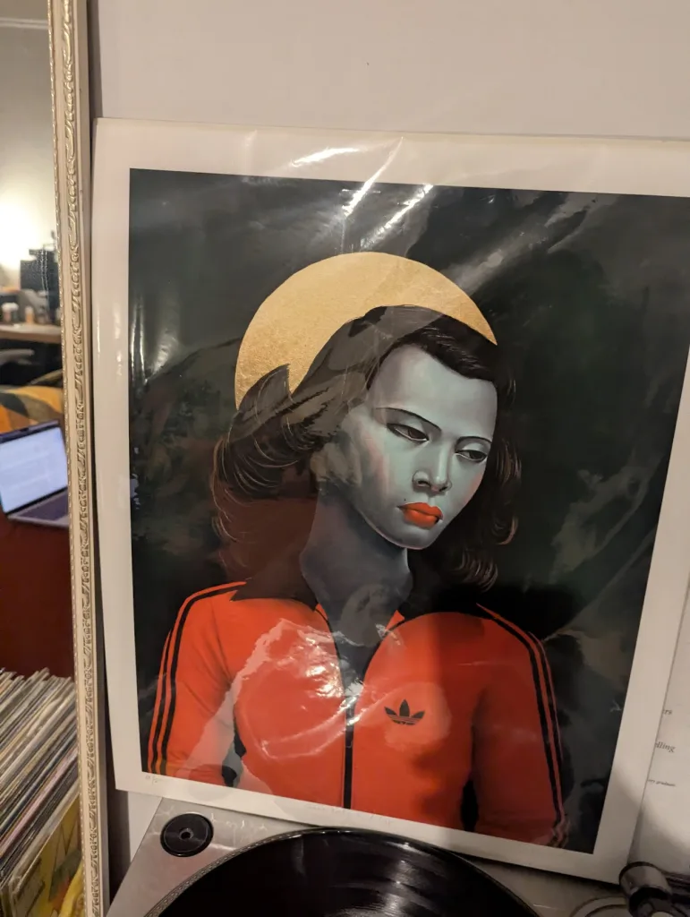Woman with blue skin and a gold halo wearing an orange Adidas tracksuit jacket in a stylized art print propped up on a record player.
