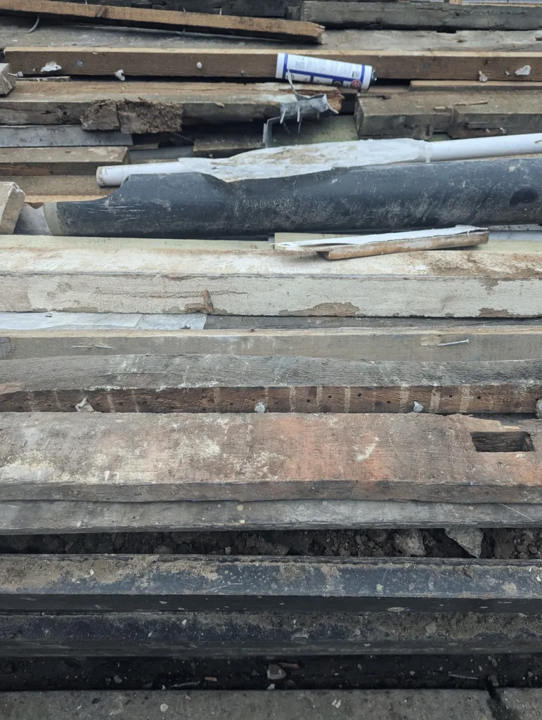 Weathered wooden planks, a large black pipe, and a tube of sealant stacked in a pile of construction debris.