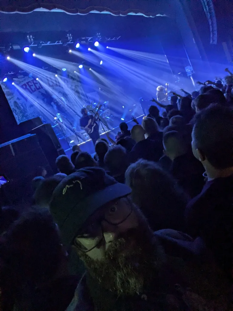A bearded man in a cap takes a selfie in a crowded concert venue with blue stage lights beaming over the band and audience.