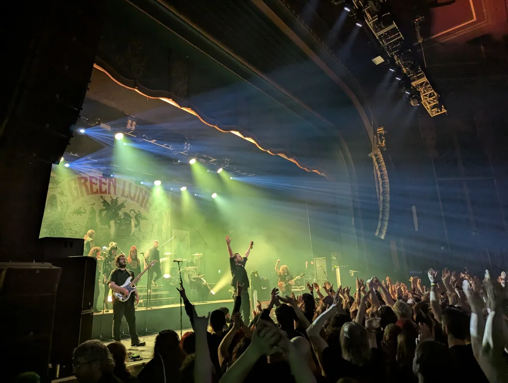 Band Green Lung performs on a theater stage under green spotlights. The singer and crowd raise their arms while light beams cut through the haze.