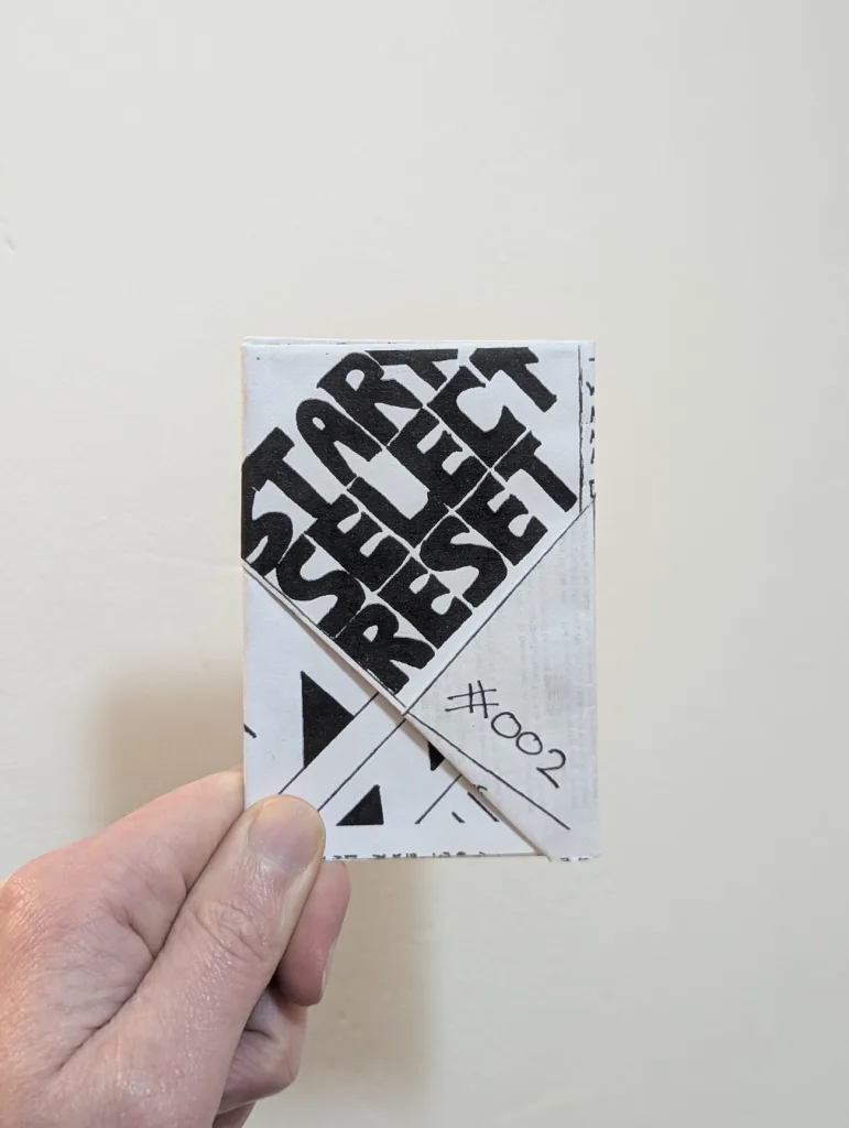 Featured image for Start Select Reset #002 - a hand holding a small folded paper zine with bold black text.