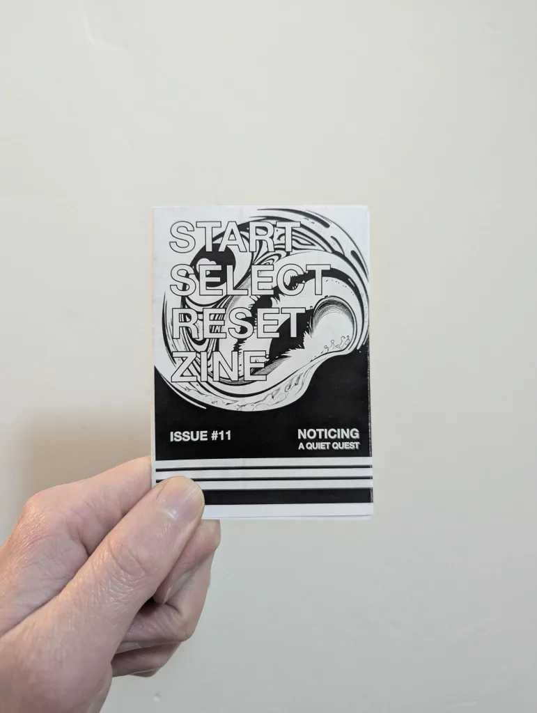 Featured image for Start Select Reset Zine Issue #11 - a hand holds a zine with swirling cover art and the text Noticing: A Quiet Quest.