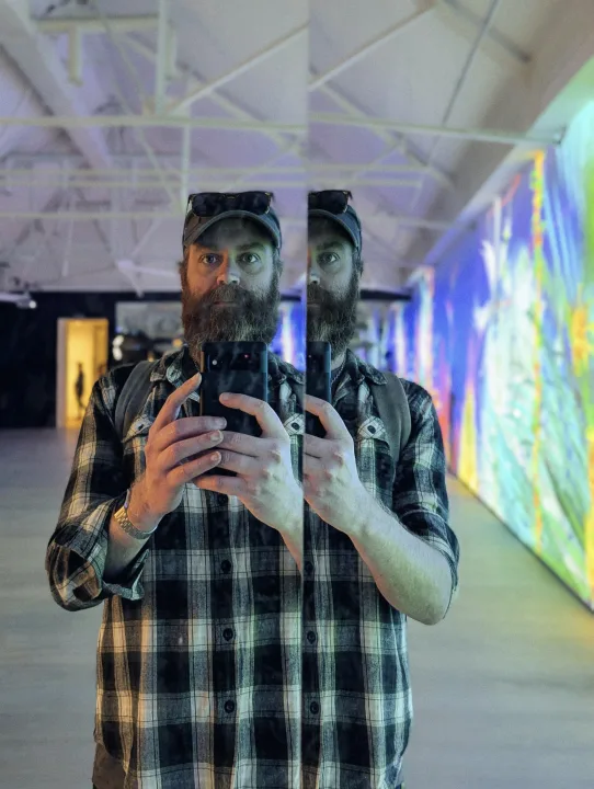Jay Springett taking a mirror selfie in a plaid shirt with a colorful light installation in the background.