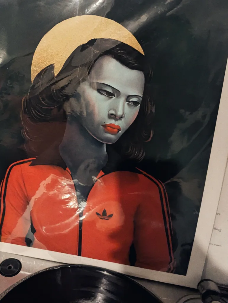 Stylized portrait of a woman with a gold halo and red lips wearing a red Adidas tracksuit jacket, propped up behind a vinyl turntable.