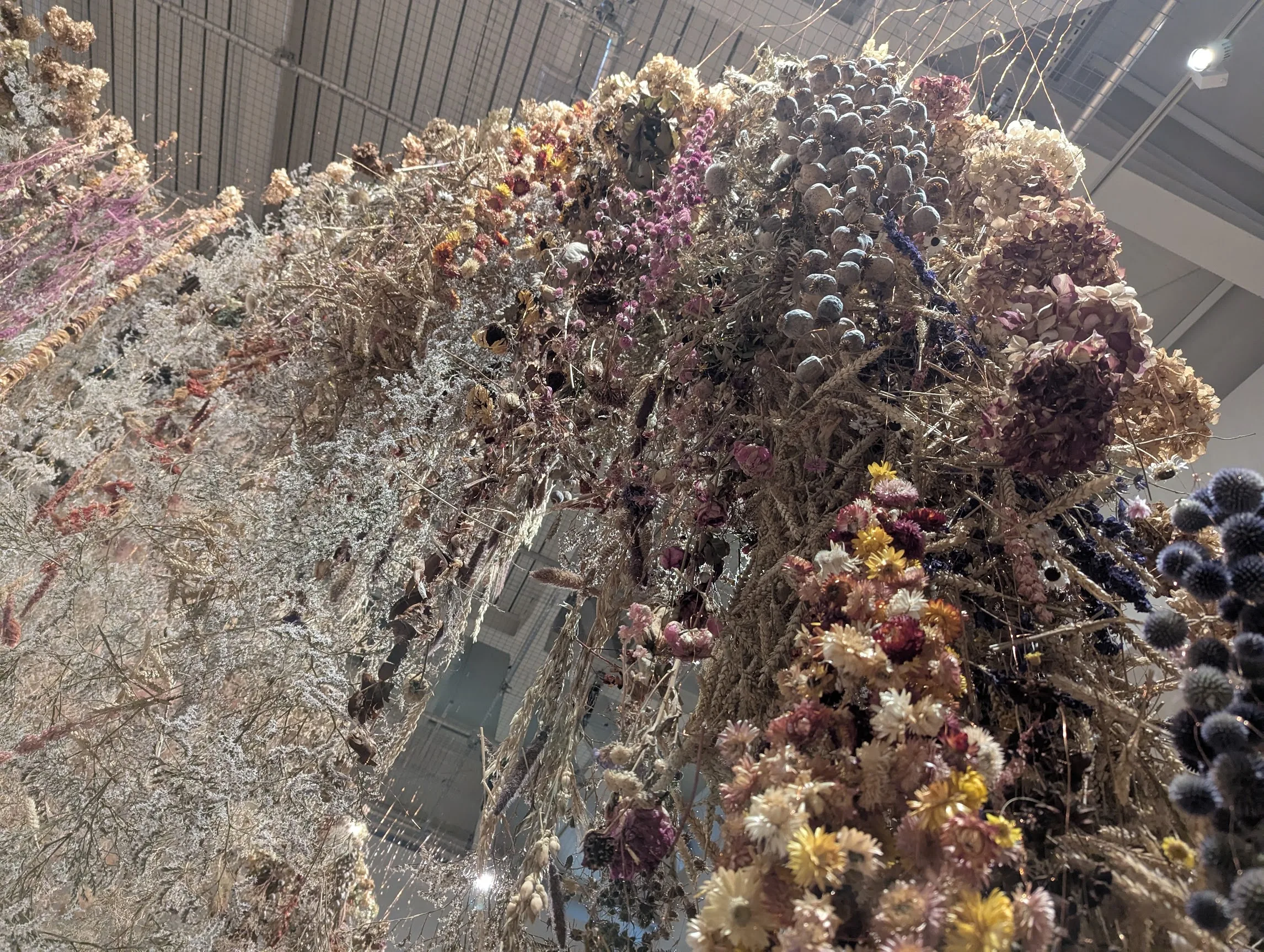 A large hanging installation of dried wildflowers, poppy pods, and thistles in muted tones against a high industrial ceiling.