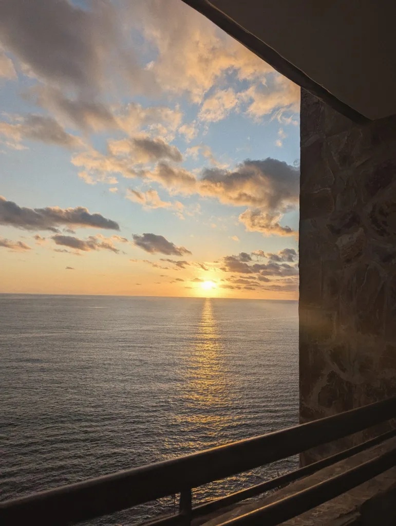 Golden sunset over the sea from a balcony, with sunlight reflecting on the water under orange-tinted clouds and framed by a stone wall.