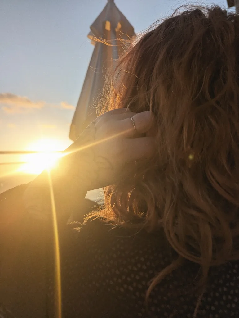 Bright sunset flare beside a person with wavy brown hair seen from behind, their hand touching their head near a patio umbrella.