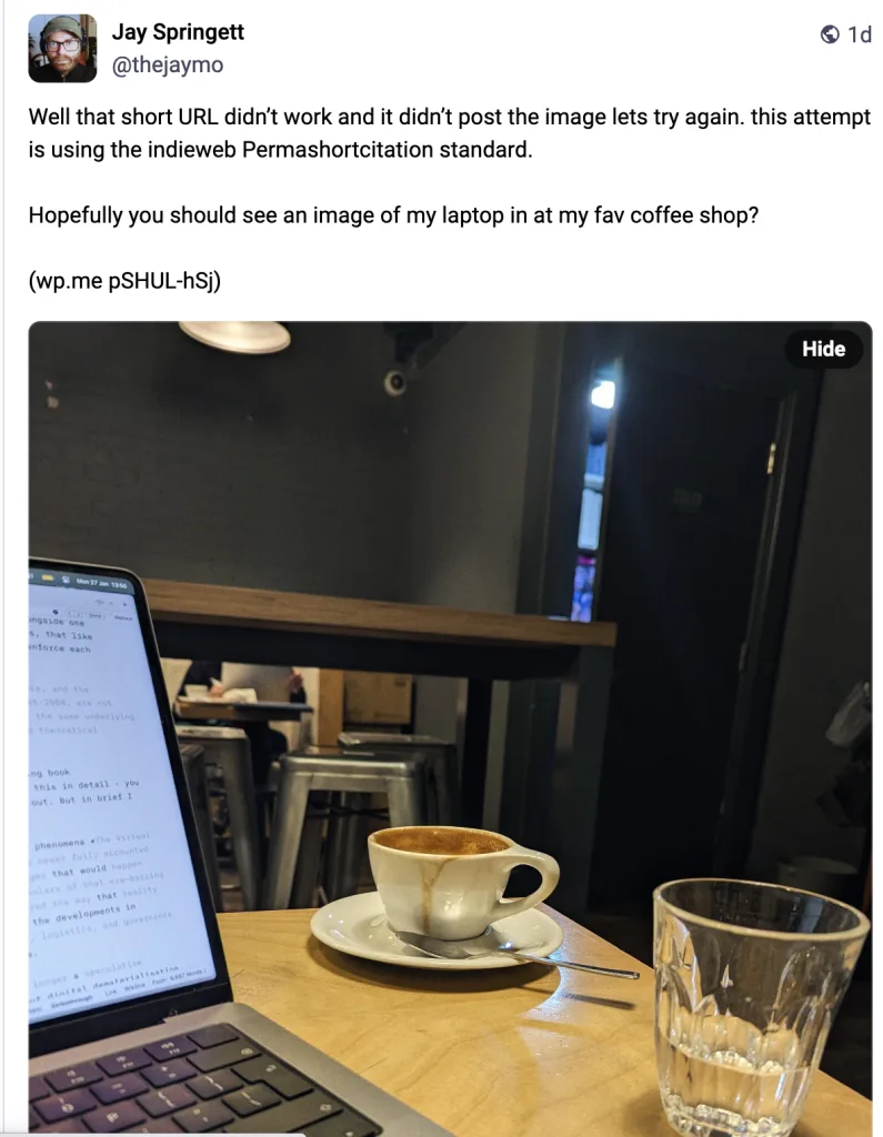 Screenshot of a Mastodon post by Jay Springett (@thejaymo) about testing the indieweb Permashortcitation standard. The post includes a photo of a laptop showing a text document next to a coffee cup and a glass of water on a wooden cafe table. Text: "Hopefully you should see an image of my laptop in at my fav coffee shop? (wp.me pSHUL-hSj)