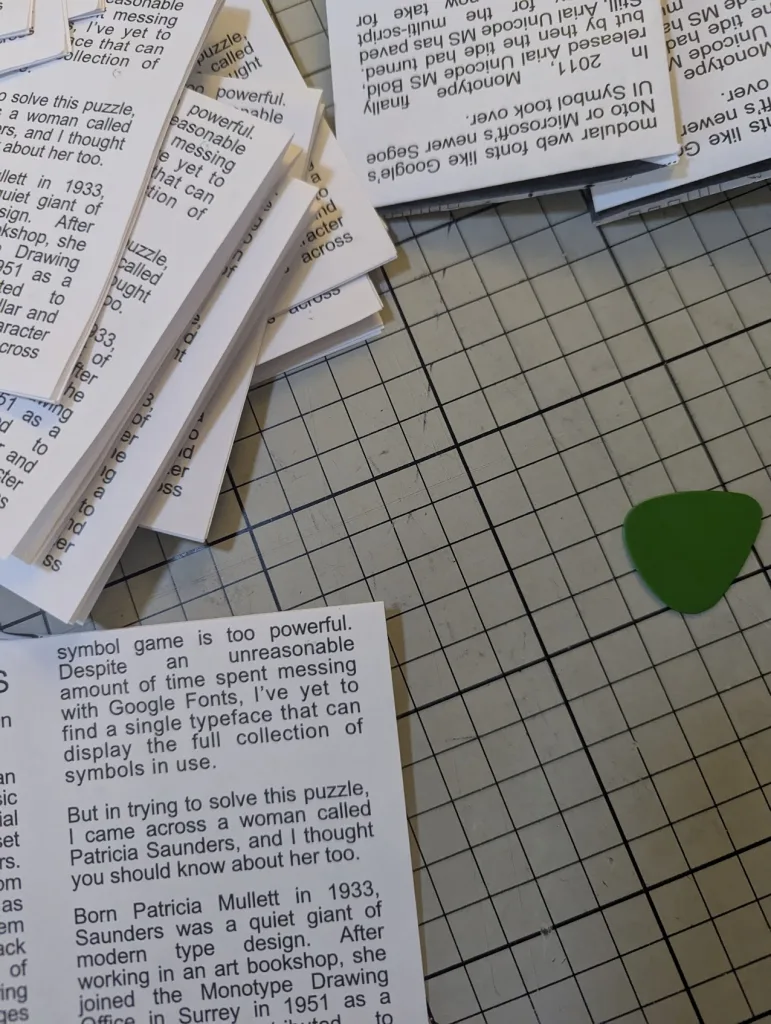 Scattered zines about typeface design and Patricia Saunders on a grey gridded cutting mat next to a green guitar pick.