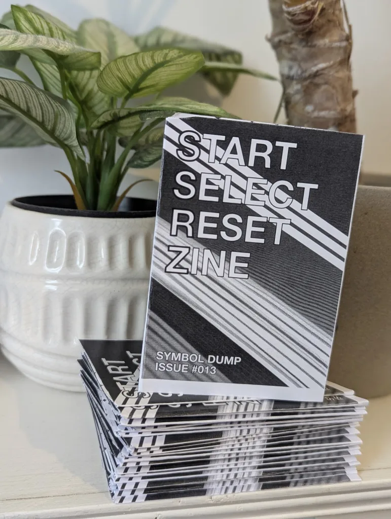 A stack of black and white Start Select Reset Zines, Issue #013 Symbol Dump, leaning against a white potted houseplant.