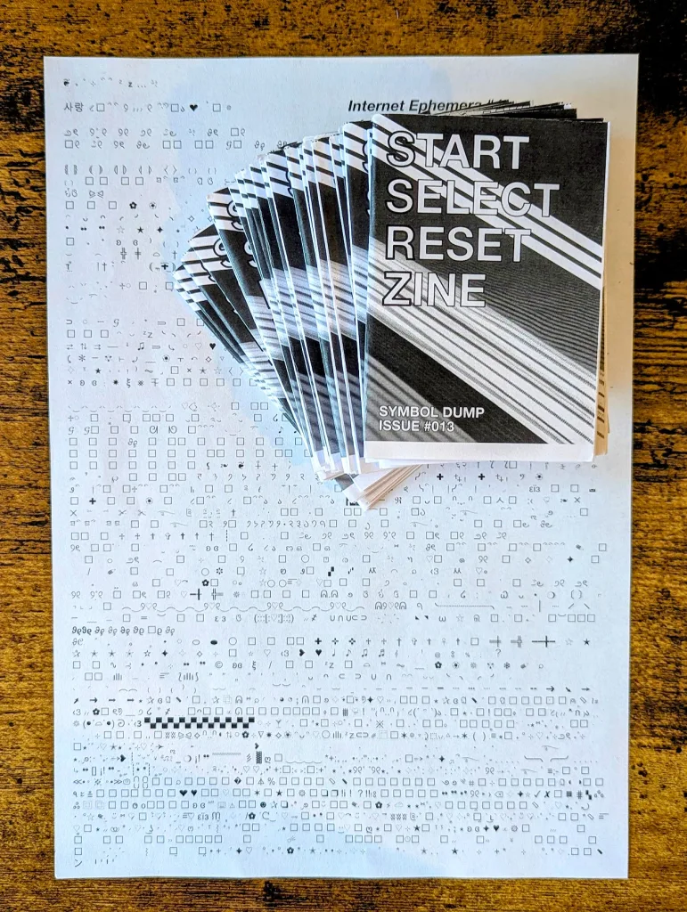 Fanned stack of START SELECT RESET ZINE Issue #013 resting on a sheet of paper covered in symbols and characters on a wooden surface.