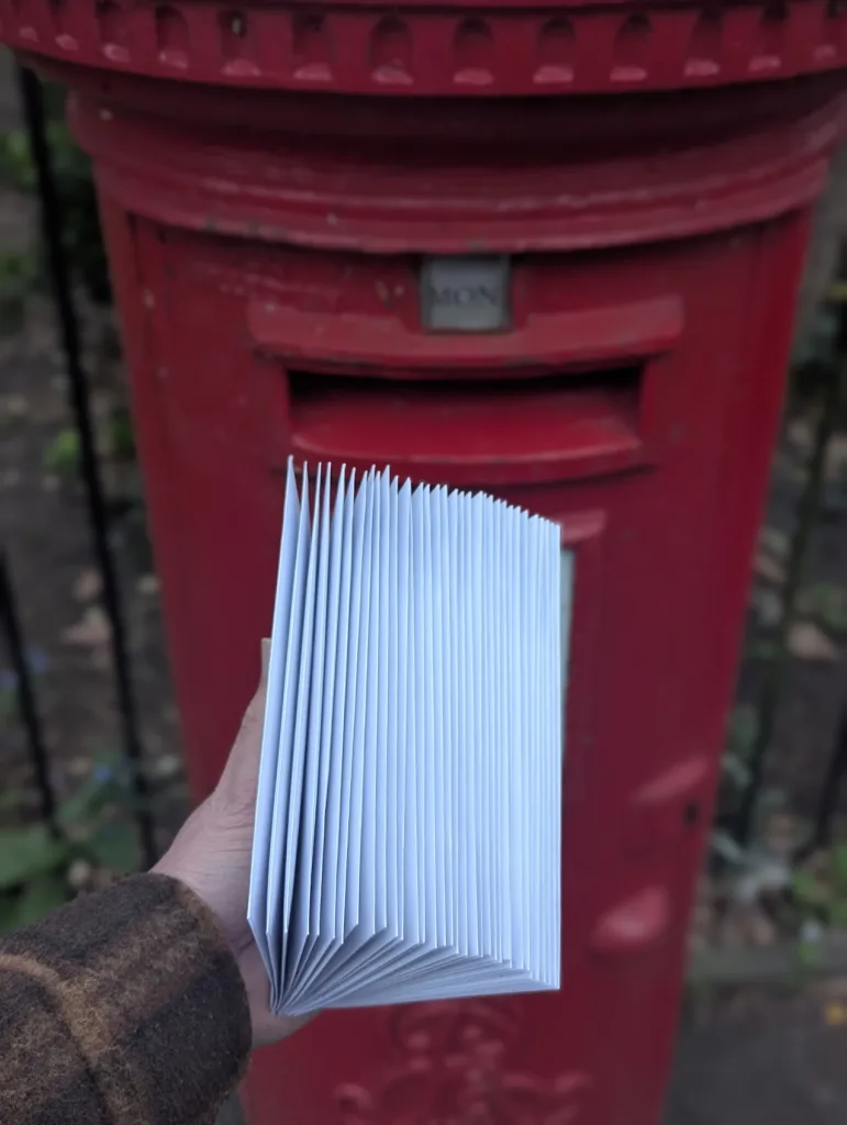 A hand holding a thick stack of white envelopes fanned out in front of a red British pillar postbox.