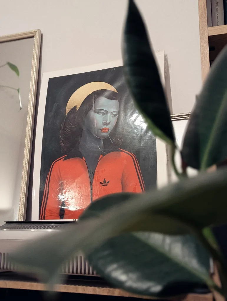 A framed print of a blue-toned woman with a gold halo wearing an orange Adidas tracksuit, viewed behind out-of-focus green houseplant leaves.