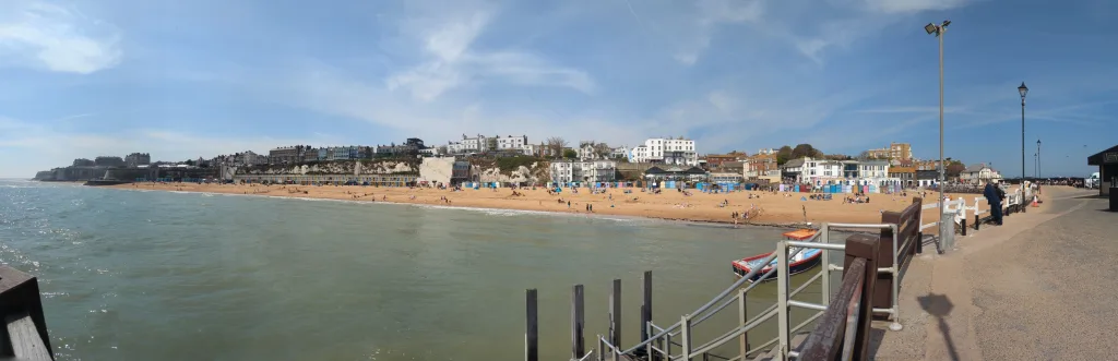 Panoramic view of a sandy beach with colorful huts and white coastal buildings along the cliffside under a bright blue sky.