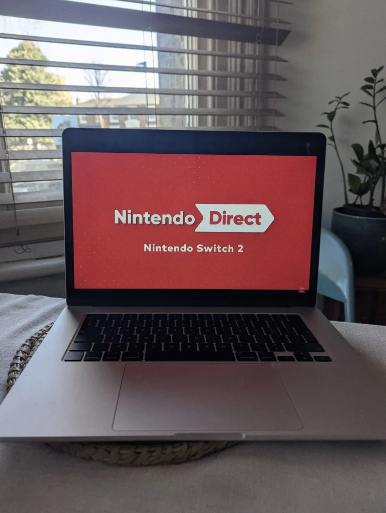 A laptop on a table displaying a Nintendo Direct logo and Nintendo Switch 2 text on a red screen, in front of a window with blinds.