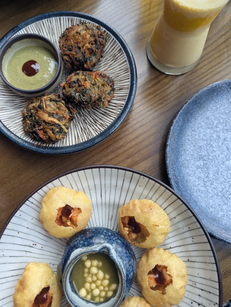 Three vegetable pakoras with green chutney and a plate of pani puri on a wooden table alongside a layered mango lassi.