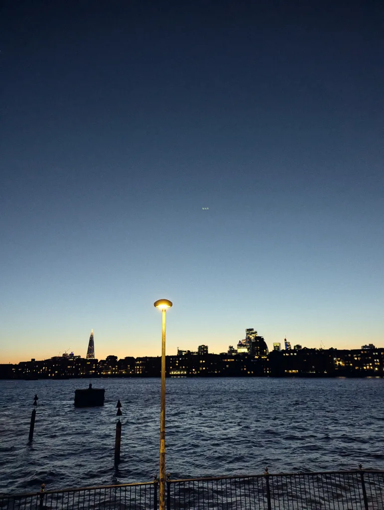 London skyline at dusk across the River Thames, featuring the Shard and a glowing yellow street lamp in the foreground.