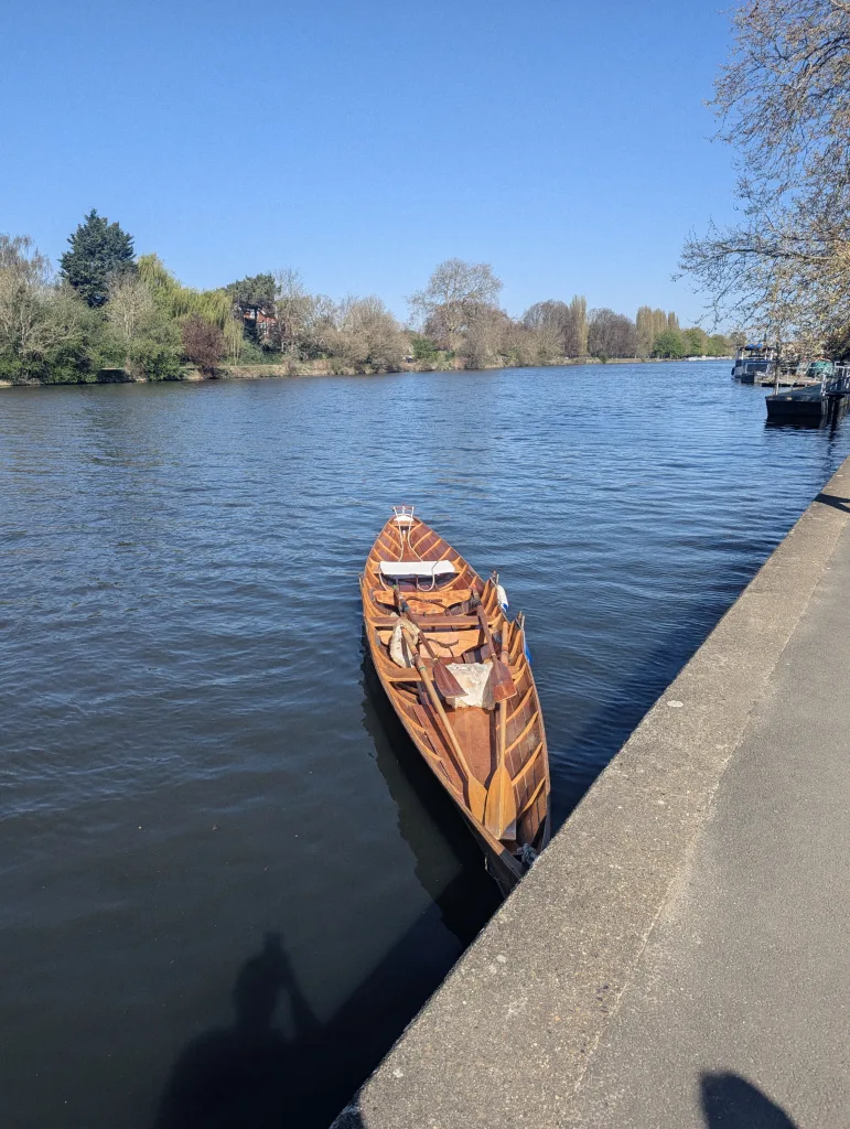 A wooden rowing skiff moored against a concrete path on a calm river under a clear blue sky, with trees lining the far bank.