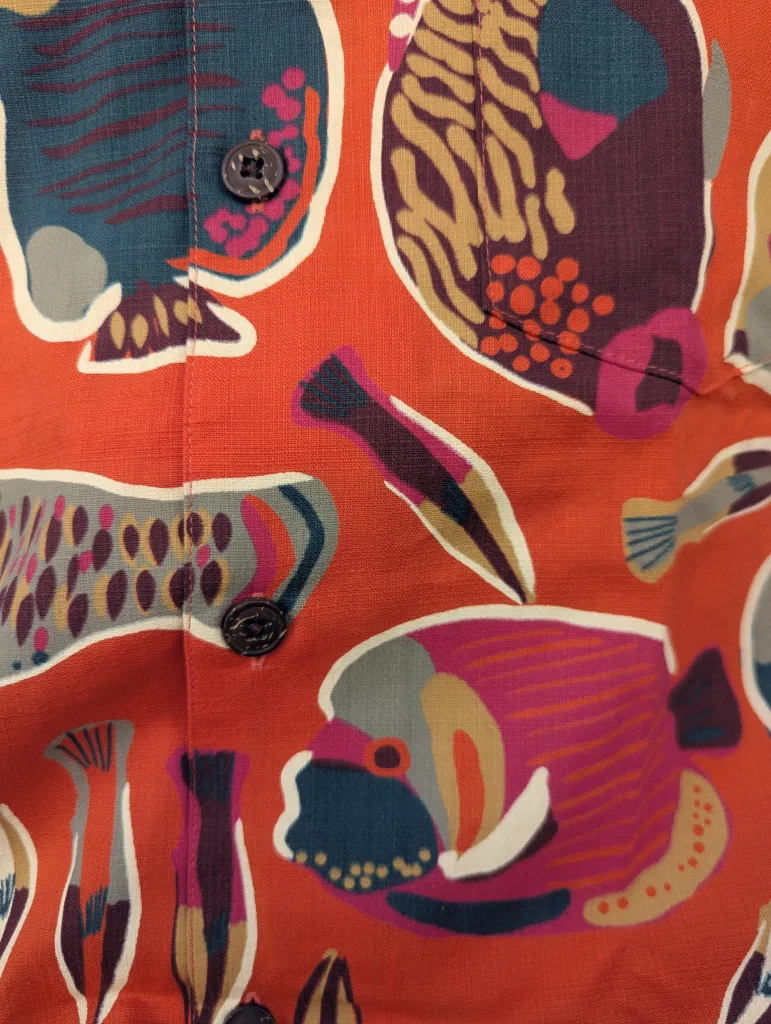 Close-up of a bright orange shirt featuring a stylized tropical fish pattern in shades of blue, pink, and tan with dark buttons.