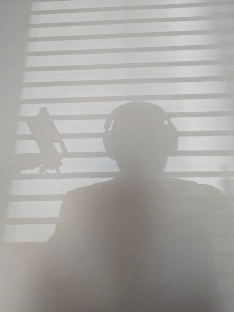 Shadow of a person wearing headphones and a boom arm microphone cast against horizontal window blinds.