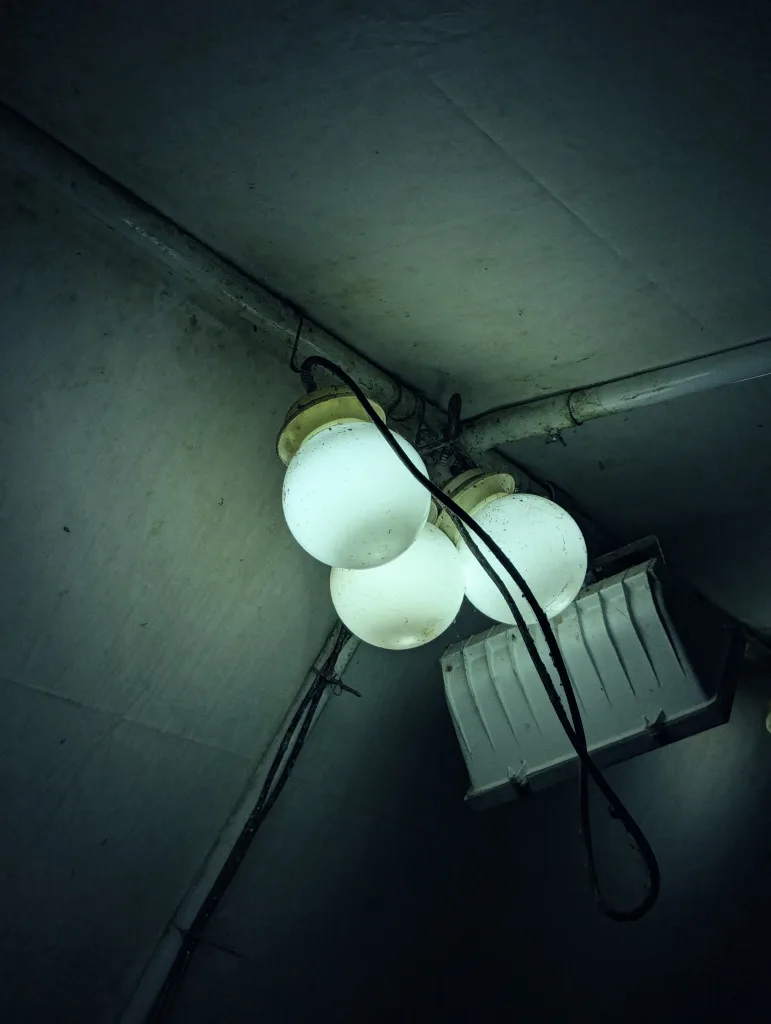 Three bright white globe lights clustered on a dark, grimy ceiling next to an industrial light fixture and a hanging black cable.
