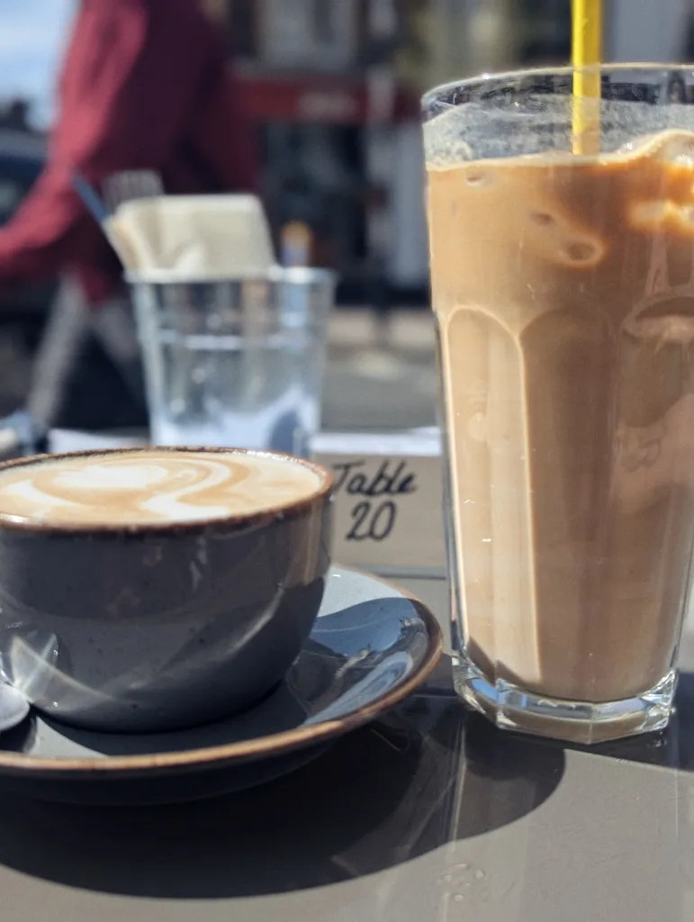 Latte art in a grey cup and a tall glass of iced coffee with a yellow straw on a sunlit cafe table next to a Table 20 marker.
