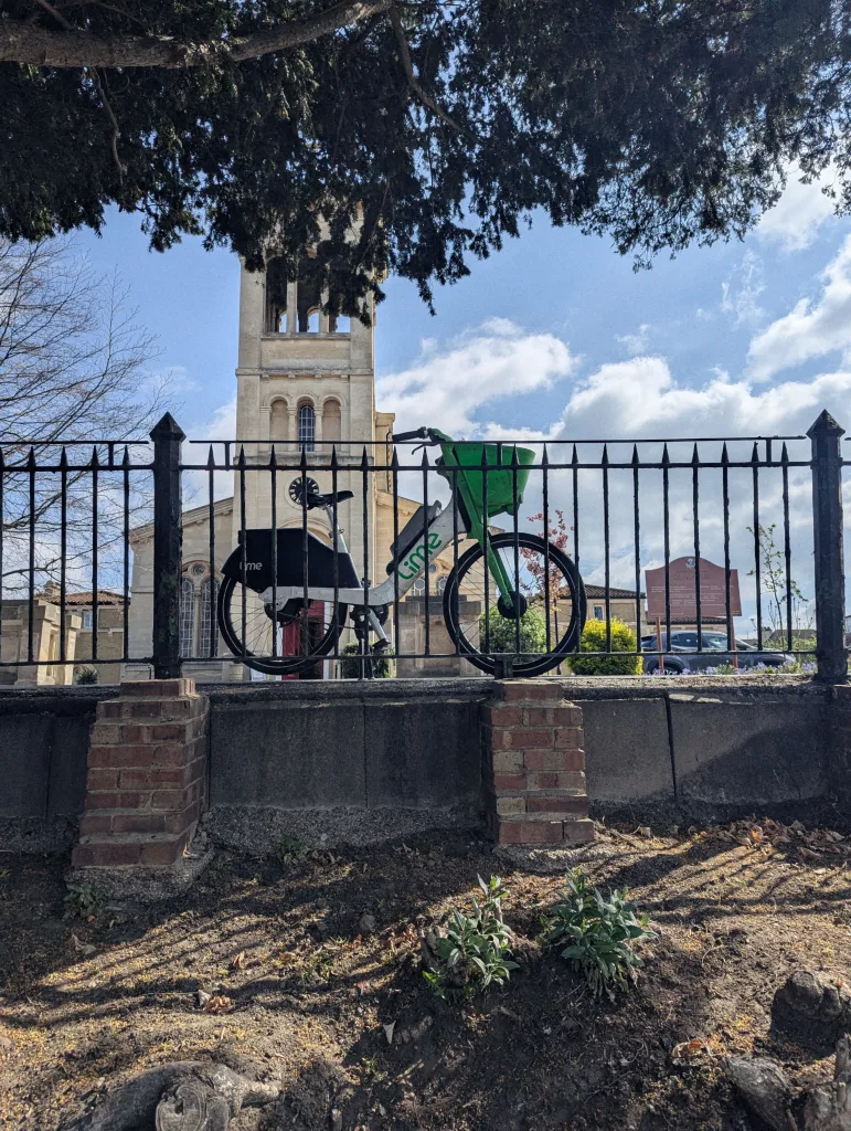 A Lime e-bike balanced on a stone wall behind a spiked metal fence, with a stone church tower and blue sky in the background.