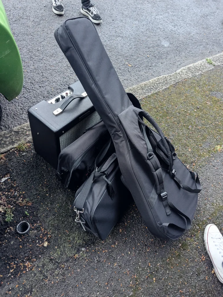 A black guitar gig bag, two gear bags, and a small amplifier sit on an asphalt street between a green car and people's feet.