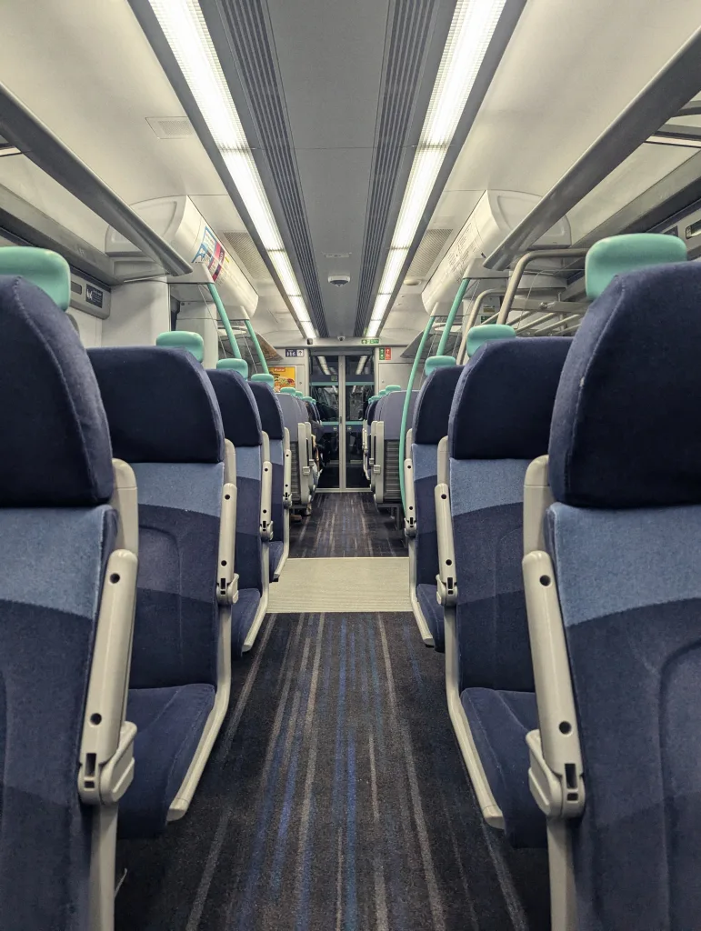 Symmetrical interior of an empty train carriage with blue seats and green headrests under bright parallel overhead strip lights.