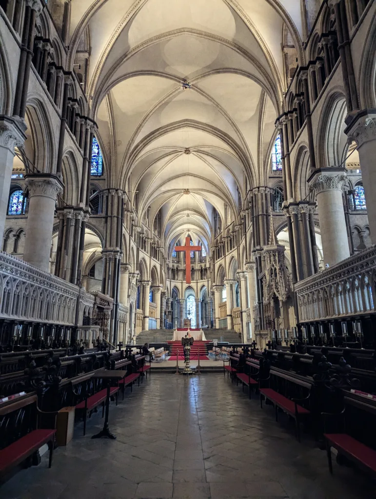 Vaulted stone arches and choir stalls inside a cathedral, with a large red cross suspended above the altar area.
