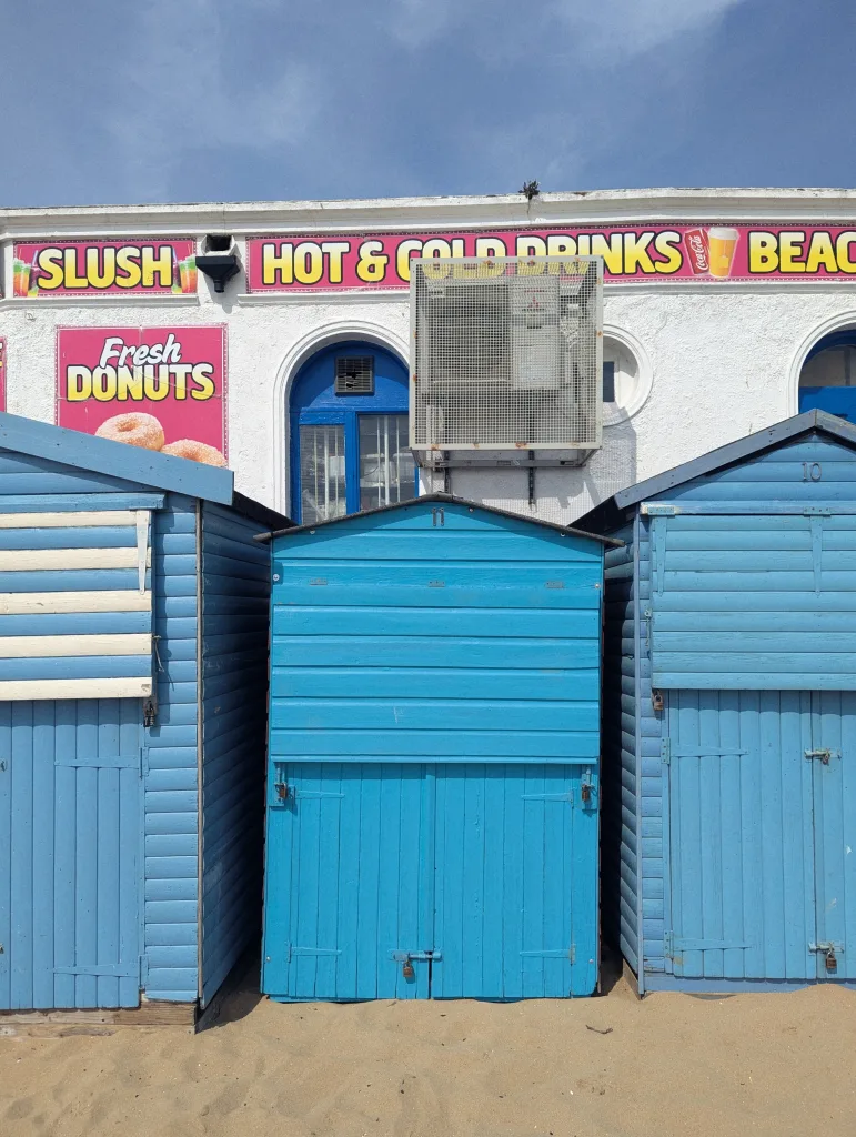 Three vibrant blue beach huts on sand in front of a seaside refreshment stall with signs for donuts and hot and cold drinks.