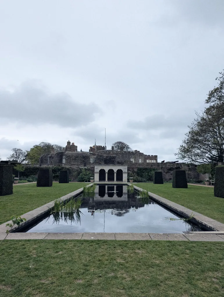 A rectangular reflecting pool centered in a formal garden leading towards a stone pavilion and the battlements of a castle under a grey sky.