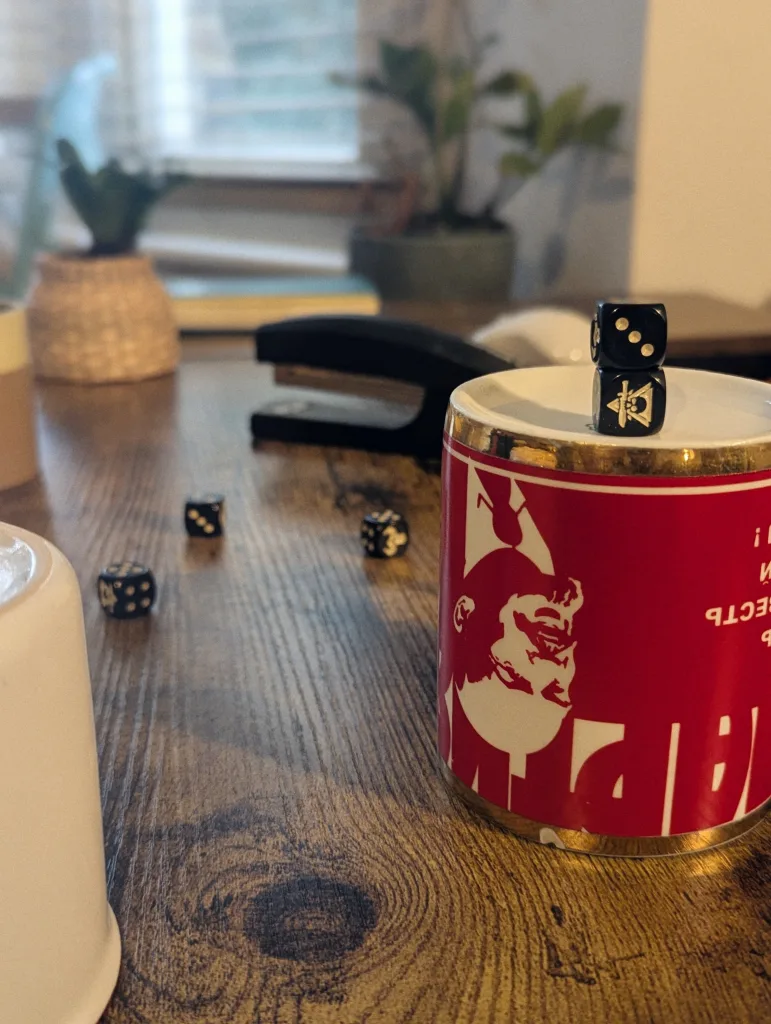 Two black dice stacked on a red mug featuring a white graphic portrait. More dice, a stapler, and plants are on the wooden desk in the background.