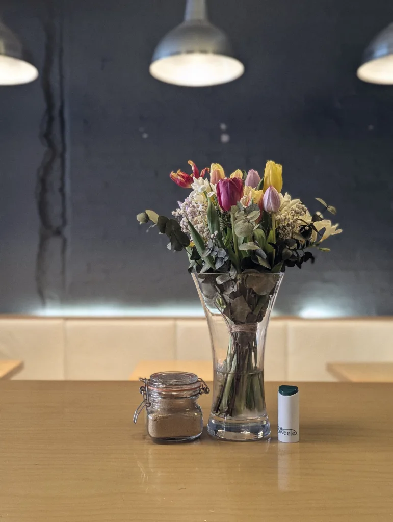 A bouquet of tulips in a glass vase, a small sugar jar, and a Sweetex dispenser on a wooden cafe table beneath industrial pendant lights.