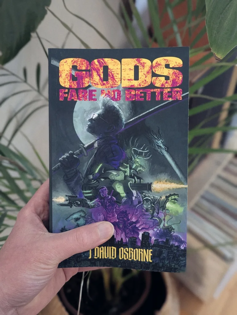 A hand holds Gods Fare No Better by J. David Osborne. The book has a neon sci-fi cover and is held in front of blurred green houseplants.