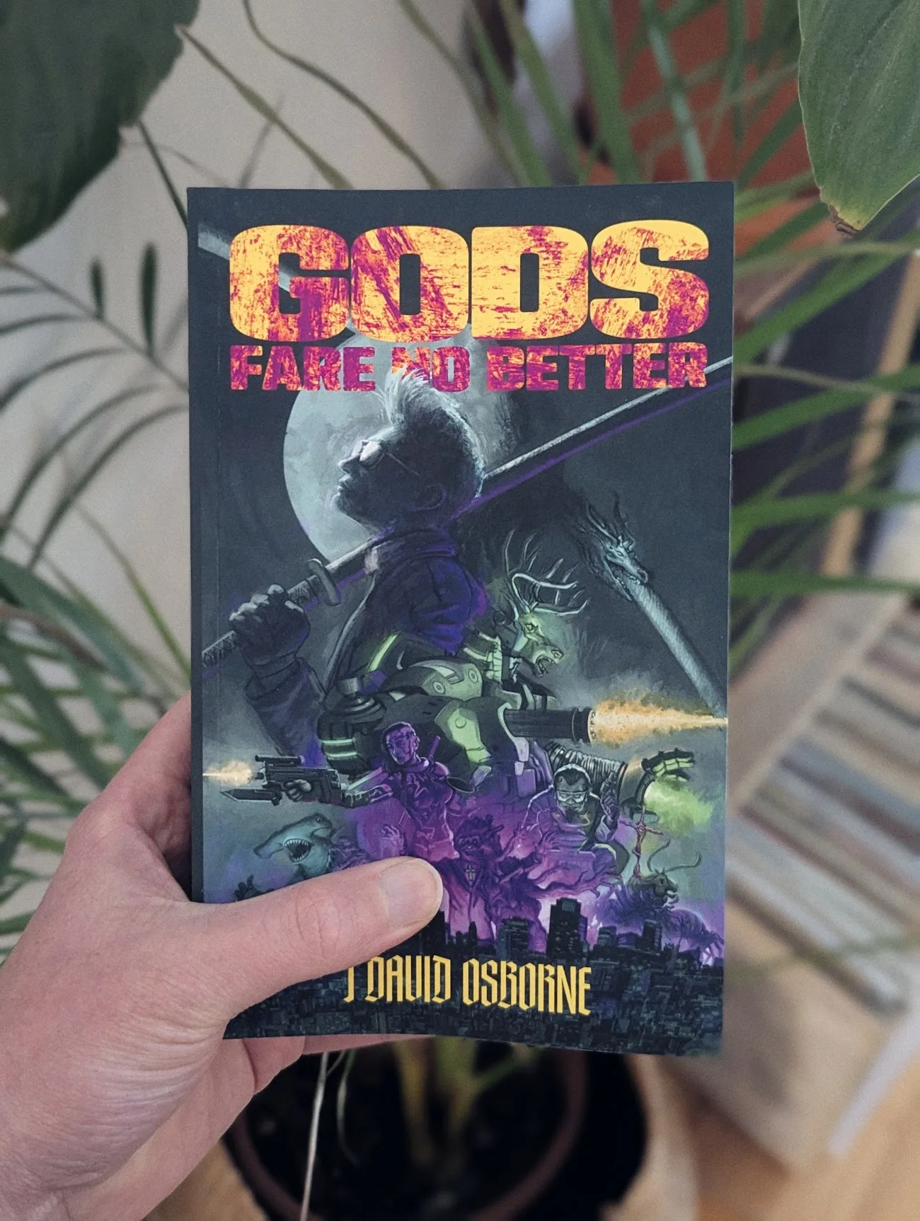 A hand holds the book Gods Fare No Better by J. David Osborne. The sci-fi cover features a man with a katana and a dragon against indoor greenery.