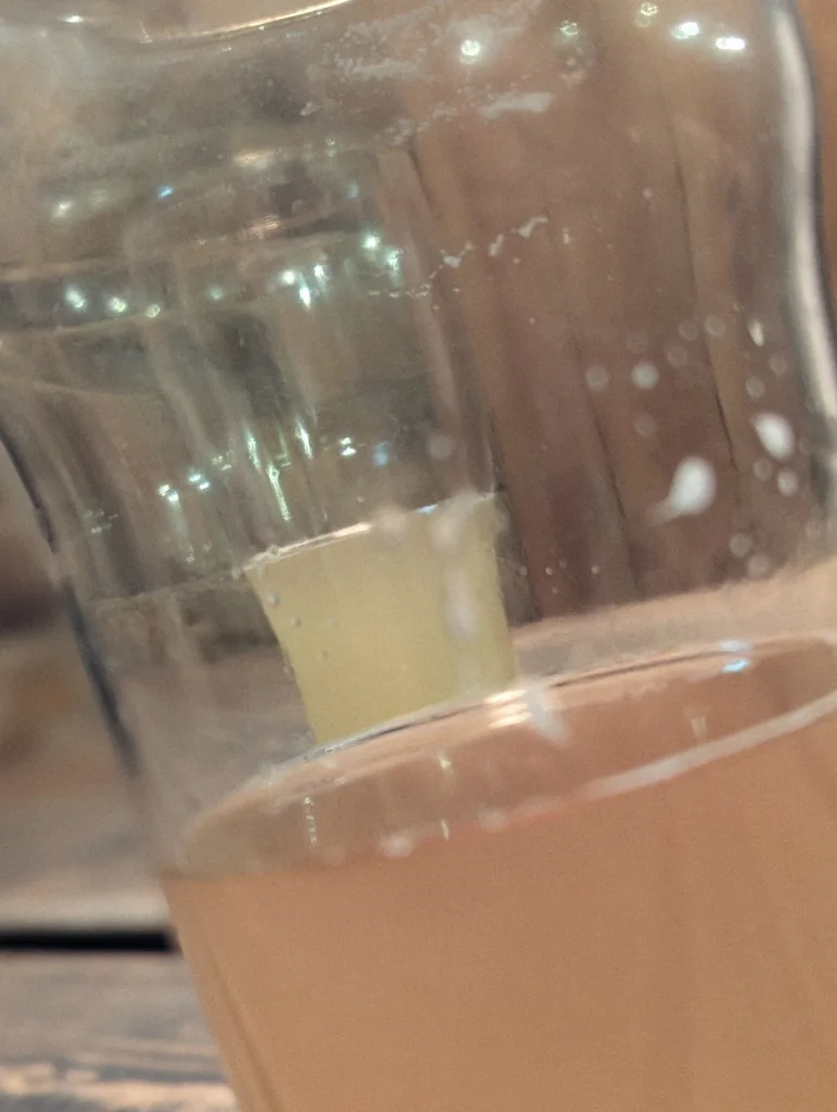 Close-up of a hazy amber liquid in a glass, with white foam lacing and bubbles visible against the clear sides.