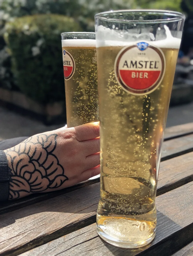 Two tall glasses of Amstel lager on an outdoor wooden table, one held by a person with a floral hand tattoo.