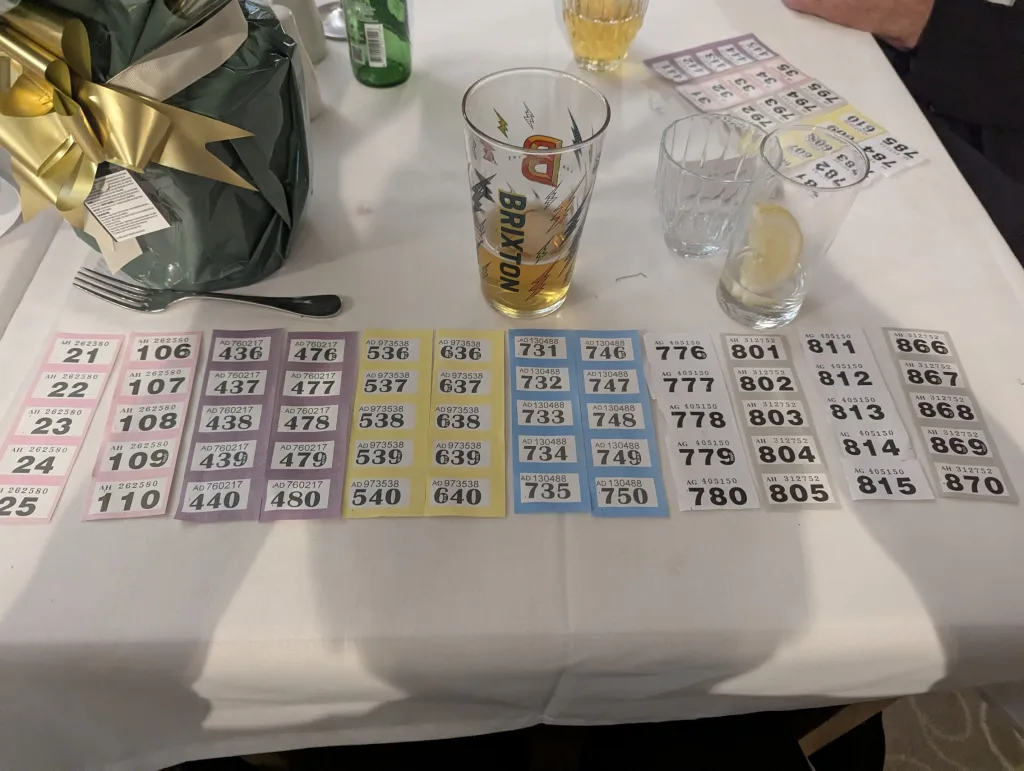 Rows of colorful raffle tickets on a white tablecloth beside a Brixton pint glass and a gift wrapped in green paper with a large gold bow.