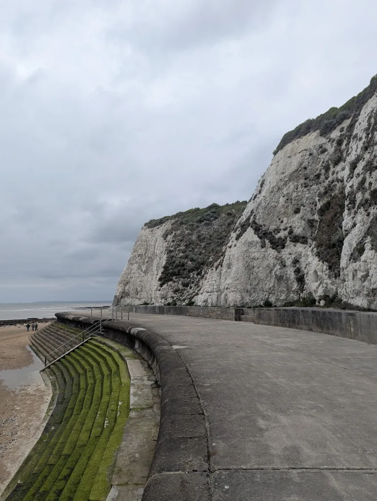 Mossy green steps lead from a concrete coastal path towards a sandy beach, bordered by towering white chalk cliffs under a grey sky.