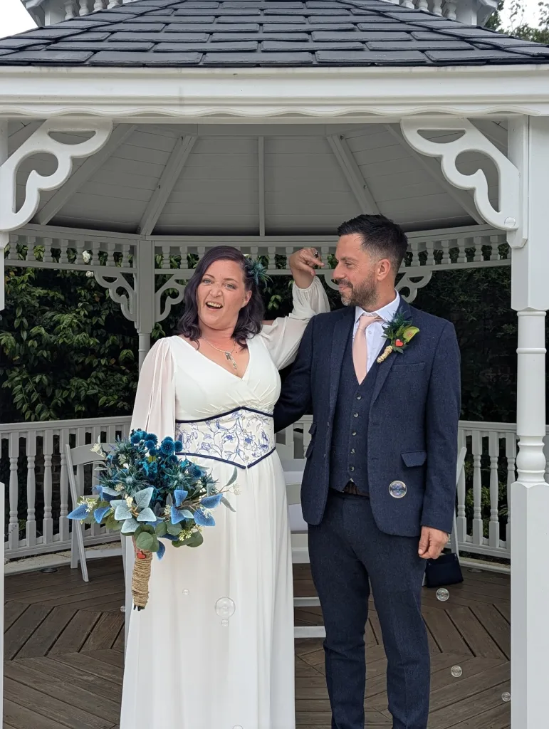 A bride in a white dress with blue floral accents and a groom in a navy suit pose playfully in a white gazebo with bubbles floating around.