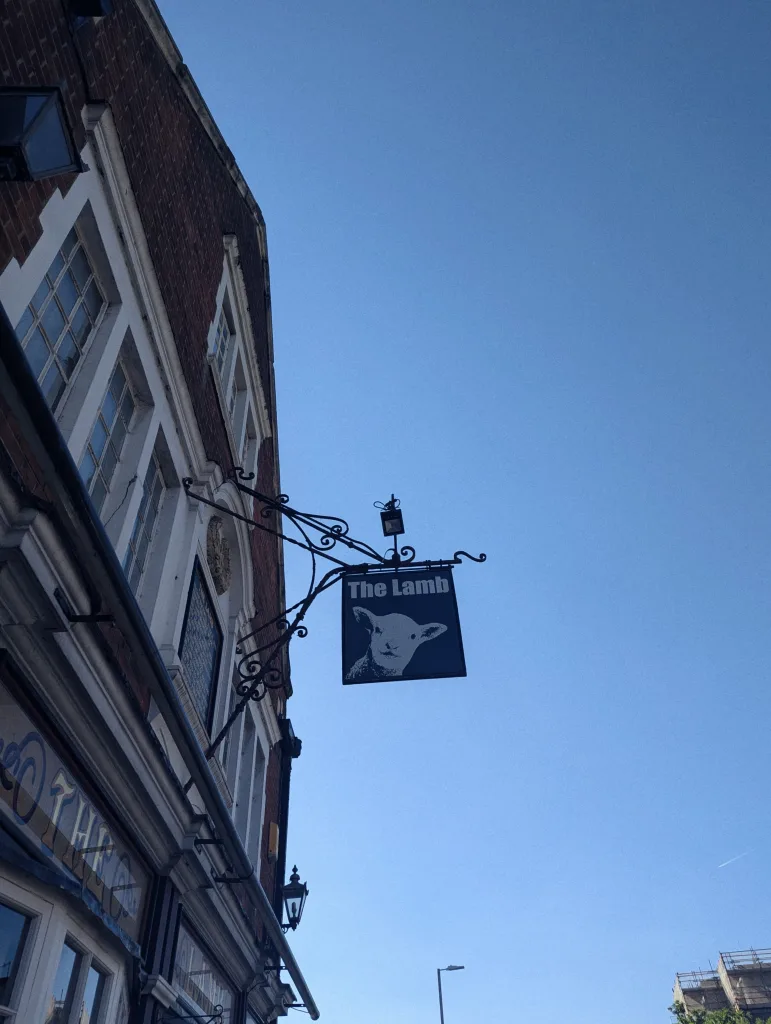 Ornate black pub sign for The Lamb featuring a lamb's head, hanging from a brick building against a clear blue sky.