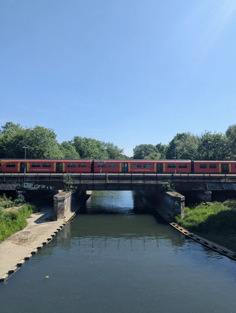 A red South Western Railway train crossing a bridge over a calm waterway, flanked by green trees under a clear blue sky.