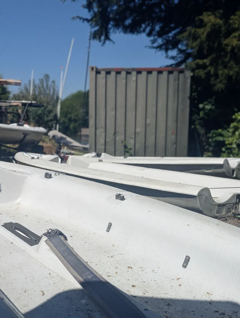 Several white sailing dinghy hulls stored on land before a grey shipping container and green trees under a clear blue sky.