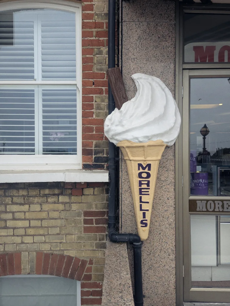 A 3D ice cream cone sign with a flake and MORELLIS text, mounted on a building wall next to a white shuttered window.