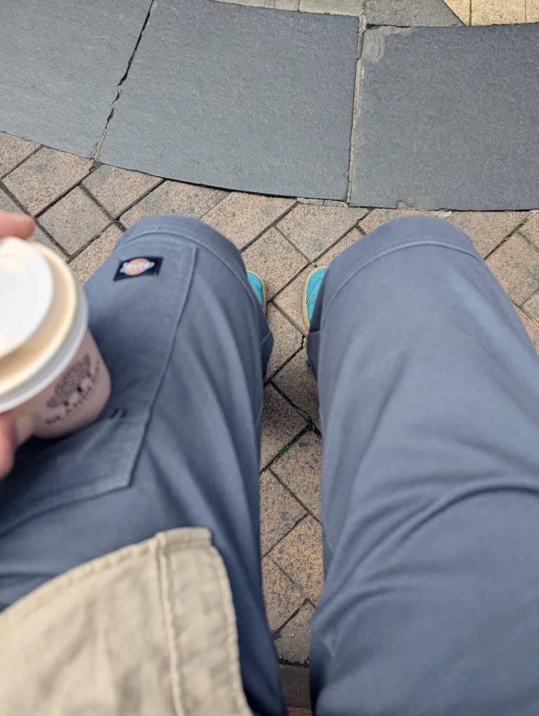 POV shot looking down at grey Dickies trousers and bright teal shoes, holding a takeaway coffee cup over brick and stone paving.