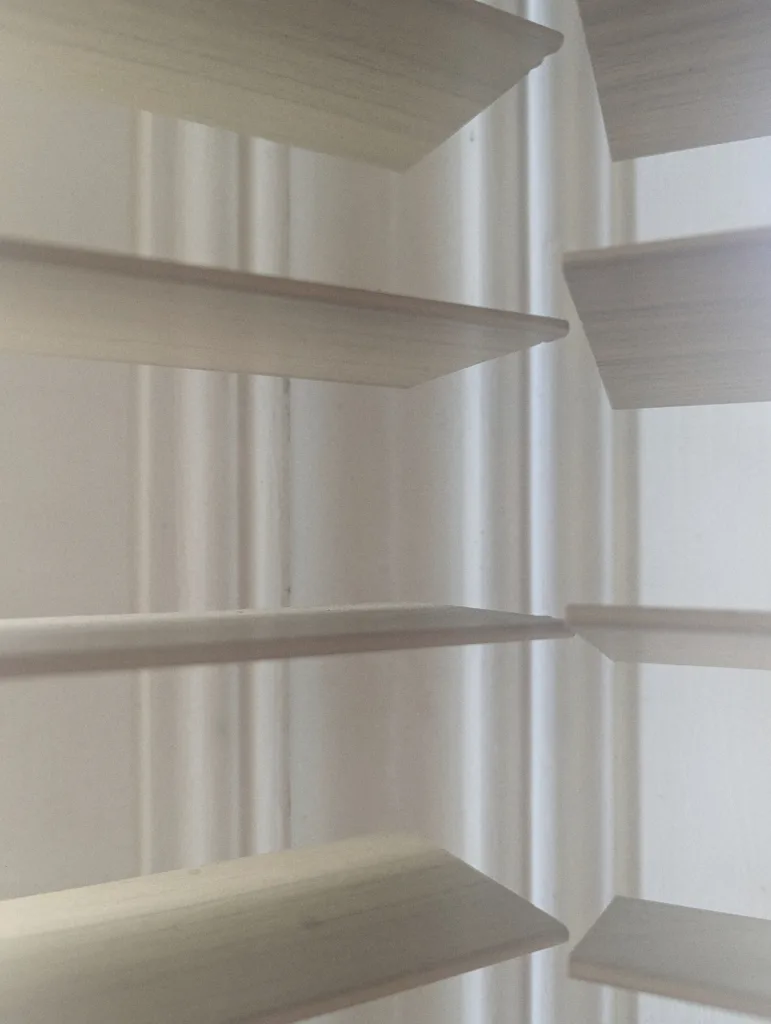 White wooden venetian blind slats angled open against a white window frame in soft light.
