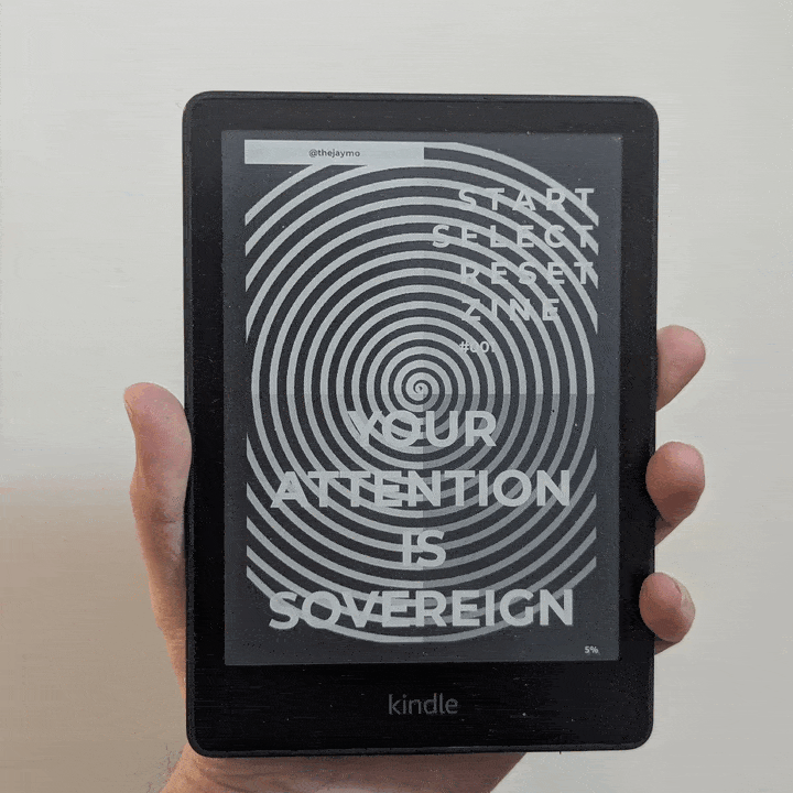 A hand holding a Kindle displaying the cover of Start Select Reset Zine #001: Your Attention is Sovereign over a black and white spiral graphic.