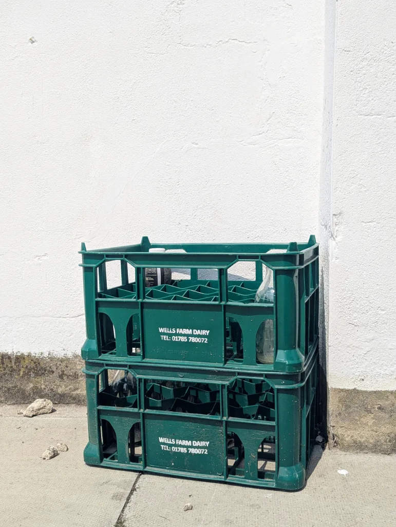Two stacked green Wells Farm Dairy plastic crates containing empty glass milk bottles against a bright, textured white wall.